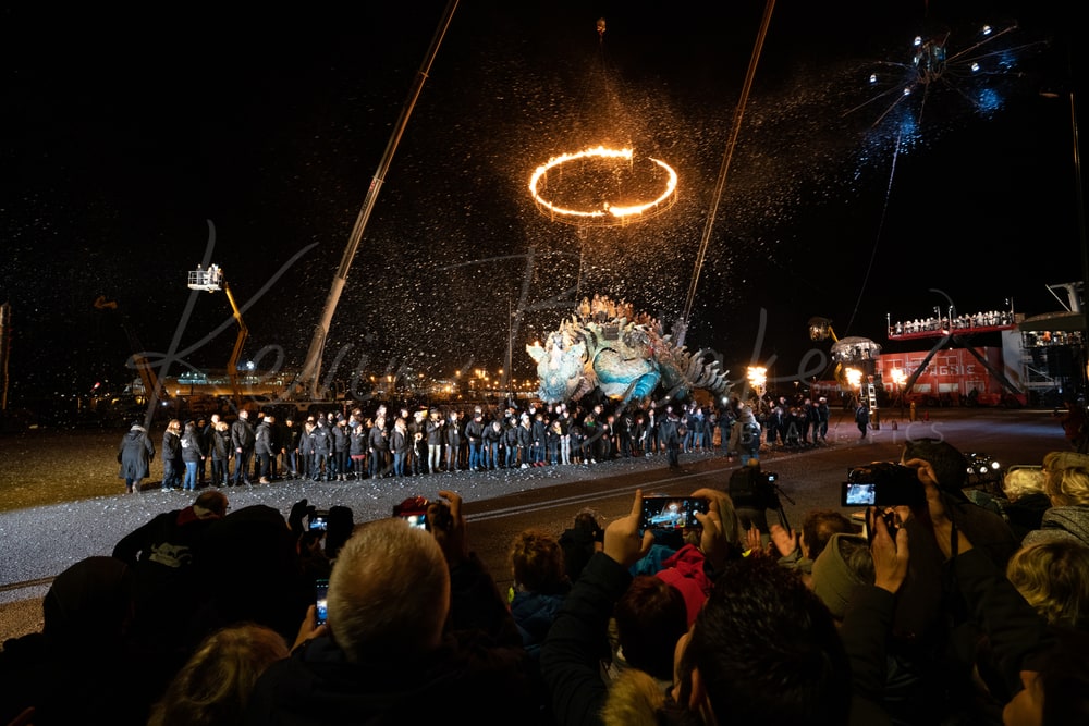 Le Dragon de Calais and performers at night beneath a ring of fire, with the crowd filming, Calais, France